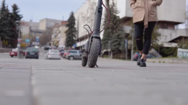 Uma jovem menina loira elegante em uma jaqueta leve e jeans preto se aproxima de uma scooter elétrica e monta-lo em um fundo de uma rua tranquila — Vídeo de Stock