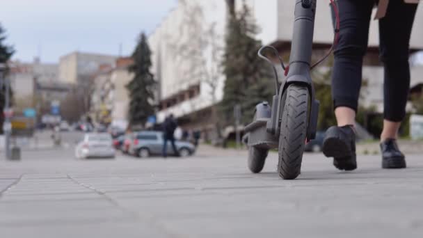 Una joven rubia con estilo en una chaqueta ligera y jeans negros se acerca a un scooter eléctrico y lo monta sobre un fondo de una calle tranquila — Vídeos de Stock