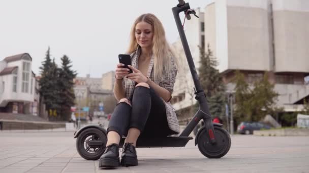 Joven chica rubia con estilo en una chaqueta ligera y jeans se sienta en un scooter eléctrico e imprime en un teléfono celular en el fondo de una calle tranquila — Vídeo de stock