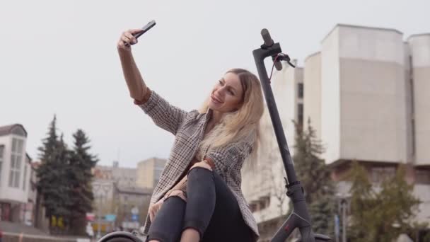 Joven chica rubia con estilo en una chaqueta ligera y jeans se sienta en un scooter eléctrico y se toma una selfie en un tranquilo fondo de calle — Vídeos de Stock