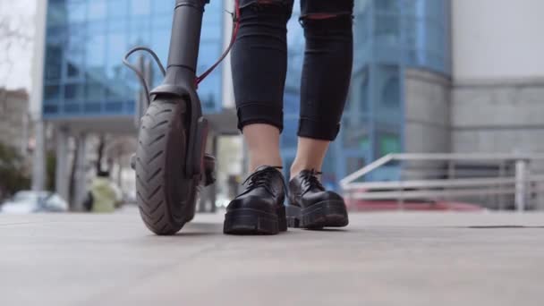 Jonge stijlvolle blonde meid in een beige trench jas en jeans staat in de buurt van een elektrische scooter in de buurt van een modern hoogbouw op de achtergrond. Voeten, schoenen en wielen — Stockvideo