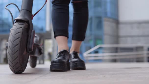 Das junge, stylische blonde Mädchen in beigem Trenchcoat und Jeans steht neben einem Elektroroller und fährt im Hintergrund an einem modernen Hochhaus vorbei. Art von Füßen, Schuhen und Rädern — Stockvideo