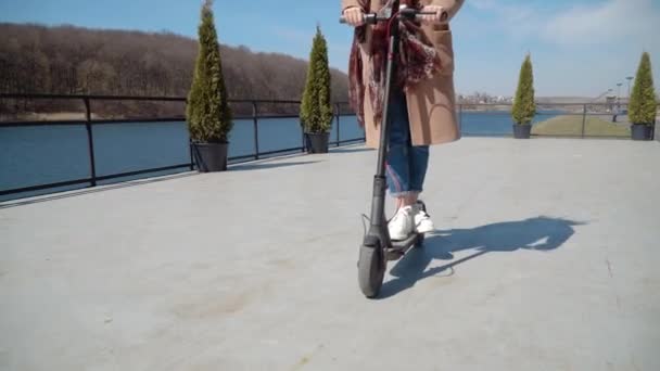 Una joven estudiante en jeans y un abrigo monta en una scooter eléctrica cerca del río. Vista de cerca del scooter y las ruedas desde el frente — Vídeos de Stock