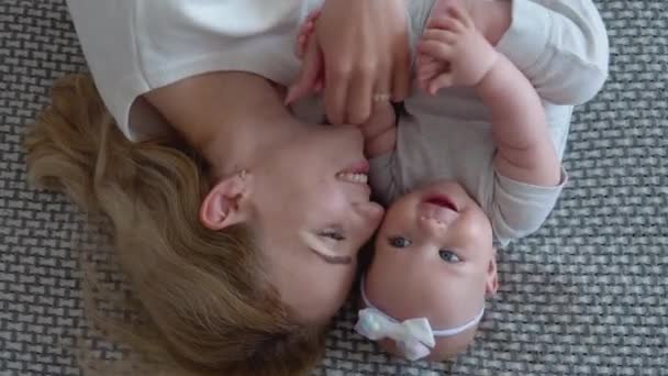 Мать и девочка лежат на кровати лицом вверх. Ребенок кусает ногу, держа ее руками. Белый и серый текстиль и одежда матери и ребенка. Вид сверху — стоковое видео