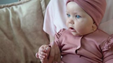 Şirin pembe takım elbiseli mavi gözlü küçük kız annesinin kucağında oturuyor. Anne ve bebek pembe elbiseli yumuşak bir kanepede kameraya dönük oturuyorlar.