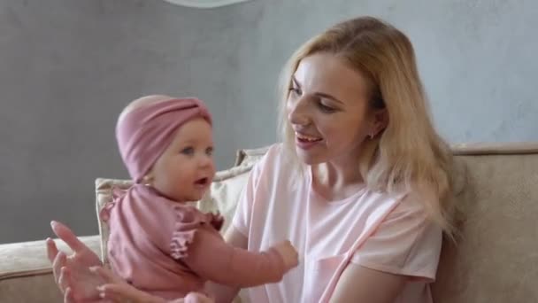 Menina bebê com olhos azuis em um terno rosa bonito brincando com a mãe em seu colo. Mãe e bebê em roupas cor-de-rosa se sentam em frente um do outro em um sofá leve macio — Vídeo de Stock