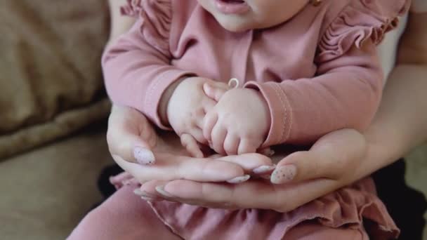 Babys mãos nas mãos das mães. Vista de perto das mãos. Menina do bebê com olhos azuis em um terno rosa bonito senta-se no colo das mães — Vídeo de Stock