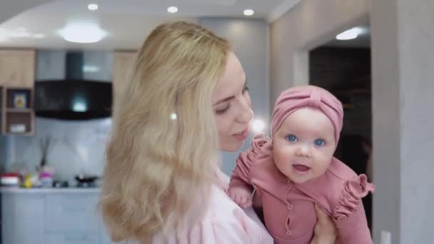 Mãe balança bebê em seus braços no fundo de um conjunto de cozinha moderna. Menina bebê com olhos azuis em um terno rosa bonito sorri e olha para a câmera — Vídeo de Stock