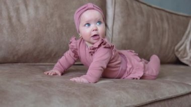 Sarı saçlı ve pembe elbiseli mavi gözlü bir bebek göbeğinde yatar ve kollarını ve bacaklarını hareket ettirir. Açık bej bir koltukta bebek