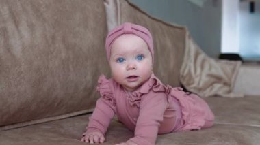 Sarı saçlı ve pembe elbiseli mavi gözlü bir kız bebek göbeğinde yatıyor ve farklı yönlerde sallanıyor. Açık bej bir koltukta bebek