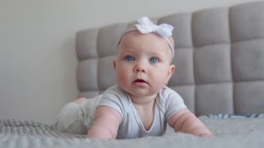 Gri elbiseli, kafasında sevimli bir fiyonk olan, yatağa uzanmış kameraya bakan küçük bir kız. Bebek bir yetişkin gördüğü için mutlu. Yaşamın ilk yılında çocuk gelişimi
