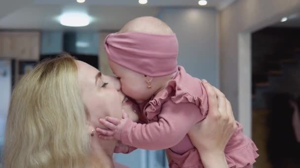 La madre gira il viso verso il bambino e fa dondolare il bambino tra le braccia sullo sfondo di un moderno set da cucina. Bambina con gli occhi azzurri in un abito rosa carino sorride e guarda la fotocamera — Video Stock