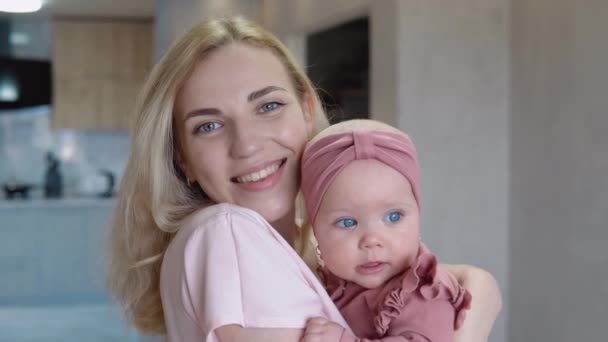 Mère et bébé fille aux yeux bleus et aux cheveux blonds regardent la caméra. La mère tient le bébé dans ses bras et embrasse sa tête — Video