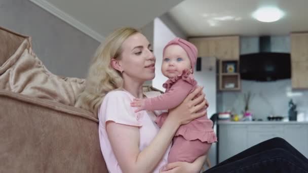 Mother and baby girl with blond hair and blue eyes in pink clothes huddle together against the backdrop of a modern kitchen set — Stock Video