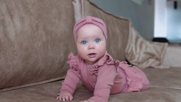 Ein kleines Mädchen mit blonden Haaren und blauen Augen in pinkfarbener Kleidung liegt auf ihrem Bauch und schwingt in verschiedene Richtungen. Baby auf hellbeigem Sofa — Stockvideo
