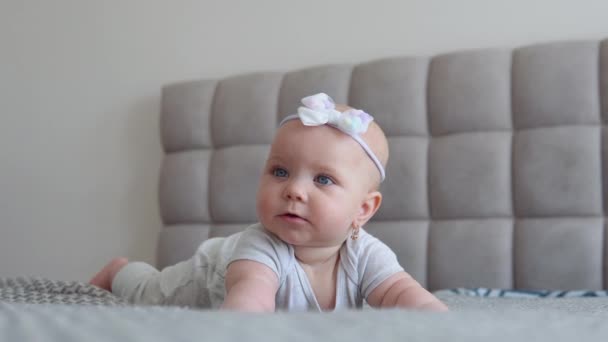 Baby Mädchen in grauer Kleidung mit einer niedlichen Schleife auf dem Kopf auf dem Bett liegend und in die Kamera blickend. Babypflege. Entwicklung des Kindes im ersten Lebensjahr — Stockvideo