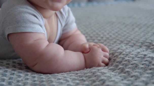 El bebé con ropa gris yace boca abajo en la cama y sonríe. Vista de cerca de la boca y las manos del bebé. Cuidado del bebé. Desarrollo infantil en el primer año de vida — Vídeo de stock