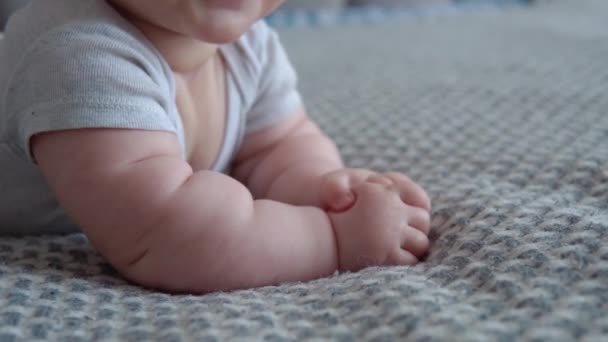 Il bambino in abiti grigi giace pancia sul letto e sorride. Vista da vicino della bocca e delle mani dei bambini. Cura del bambino. Sviluppo del bambino nel primo anno di vita — Video Stock