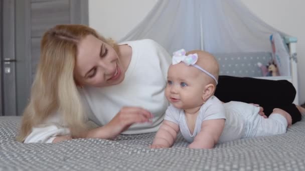 Bebê menina em roupas cinza com um arco bonito em sua cabeça encontra-se barriga para baixo na cama perto de sua mãe no fundo de um dossel com um dossel. Cuidados com o bebê. — Vídeo de Stock