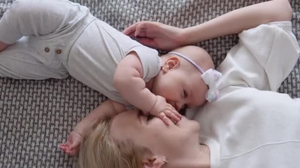 母亲和女婴脸朝上躺在床上.母亲吻着婴儿的脸。白色和灰色的纺织品和母子服装。顶部视图 — 图库视频影像