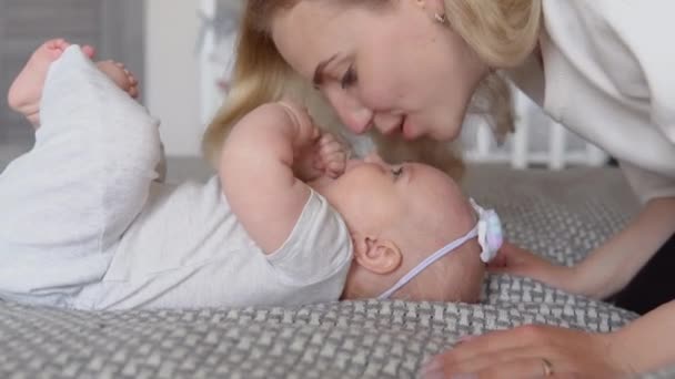 A kislány arccal felfelé fekszik az ágyon. Az anya megcsókolja a babák arcát, ahogy felette lóg. Fehér és szürke textíliák, valamint anya és gyermek ruhái. Oldalnézet — Stock videók