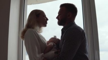 Anne ve baba pencerenin önünde durup birbirlerine bakıyorlar ve çocuk annelerinin kollarında ikisinin arasında uyurken sarılıyorlar. Babalık ve annelik. Geleneksel aile