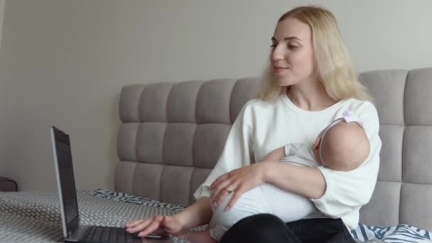 母親がラップトップで自宅で働いている間、赤ん坊の女の子は母親の腕の中で眠っています。産休に関するリモートワーク — ストック動画