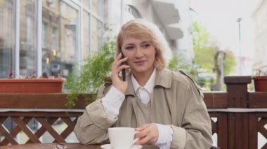 Terasta masada bir fincan kahveyle cep telefonuyla konuşan bir kadın. Trençkotlu ve beyaz bluzlu bir kadın beyaz bir kahve fincanı aldı ve masaya geri koydu.