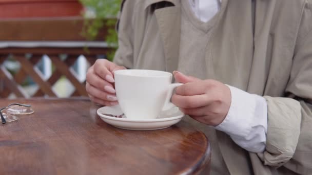 Een vrouw die 's morgens koffie drinkt op het terras. Een vrouw in een trench jas en witte blouse pakt een witte koffiekop en zet het terug op de tafel — Stockvideo