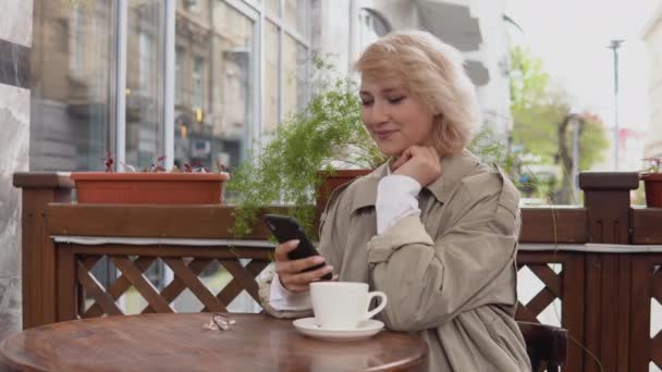 女人在阳台上的桌上用手机和一杯咖啡。一个穿着风衣和白衬衫的女人拿起一个白色的咖啡杯，放在桌上 — 图库视频影像