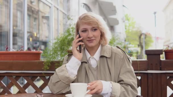Frau telefoniert mit einer Tasse Kaffee an einem Tisch auf der Terrasse. Eine Frau im Trenchcoat und weißer Bluse nimmt eine weiße Kaffeetasse und stellt sie wieder auf den Tisch. — Stockvideo