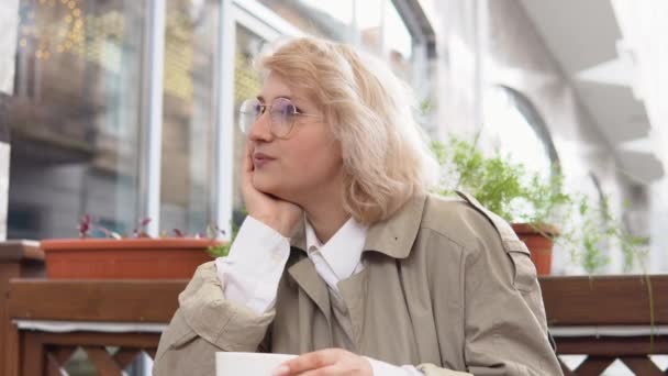 Uvolněná žena sedící se šálkem kávy u stolu na terase. Žena v plášti a bílé blůze zvedne bílý šálek kávy a položí ho zpátky na stůl. Mindfullness — Stock video