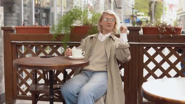 Kvinna chillar ut medan vila sitter med en kopp kaffe vid ett bord på terrassen. En kvinna i trenchcoat och vit blus plockar upp en vit kaffekopp och lägger tillbaka den på bordet. — Stockvideo
