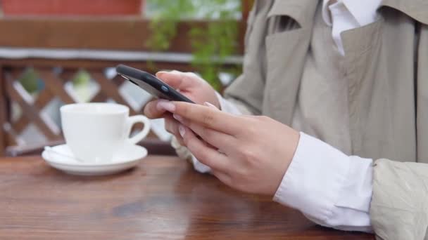 En kvinne sveiper i telefonen og sitter ved et bord med en kopp kaffe på terrassen. Nærbilde av hender med mobiltelefon. Hvit porselenskopp med bakgrunn – stockvideo