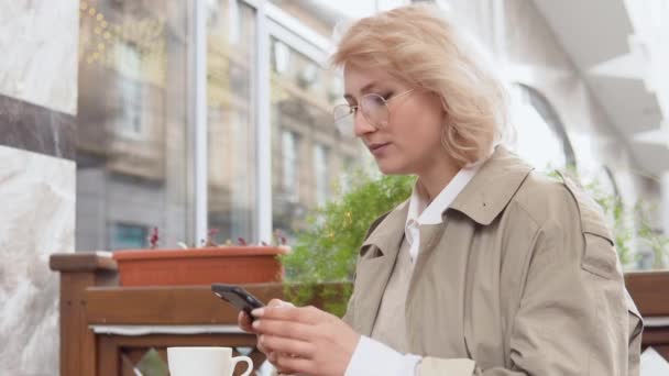 Mulher desliza no telefone sentado em uma mesa com uma xícara de café no terraço. Copo de porcelana branca em um fundo — Vídeo de Stock