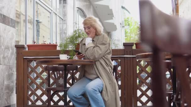 Allegro giovane donna bionda in abiti casual striscia nel telefono seduto a un tavolo con una tazza di caffè sulla terrazza — Video Stock