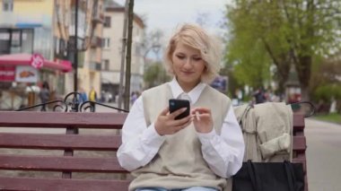 Bej yelek ve beyaz bluzlu genç sarışın iş kadını şehirdeki bir bankta oturuyor ve bir sokak kafesinin arka planındaki cep telefonunda çalışıyor.