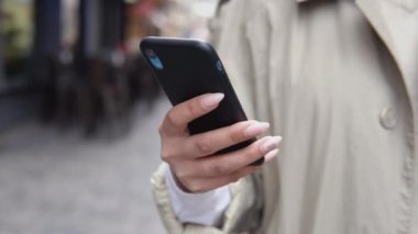 Bej renkli trençkotlu genç bir iş kadını ve sokakta duran bir cep telefonunda çalışan beyaz bir bluz. Elin yakın görüntüsü