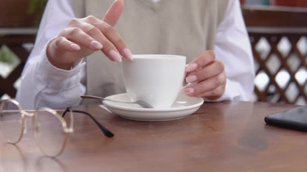 Jeune femme avec une manucure élégante dans des vêtements décontractés de couleurs beige et blanc court son doigt le long du bord d'une tasse de café en porcelaine blanche. Rituels matinaux d'une jeune femme moderne — Video