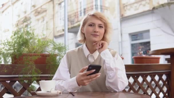 Mladá blondýnka s elegantní manikúrou v béžové vestě a bílé blůze drží mobilní telefon, zatímco sedí na letní terase v pouliční kavárně — Stock video