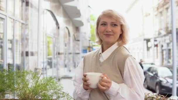 Mladá blondýna s elegantní manikúrou v béžové vestě a bílou blůzou s bílým porcelánovým šálkem s kávou, stojící na letní terase v pouliční kavárně — Stock video