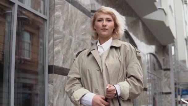 Молода блондинка в бежевому траншеї пальто і біла блузка стоїть на тлі сучасної офісної будівлі — стокове відео