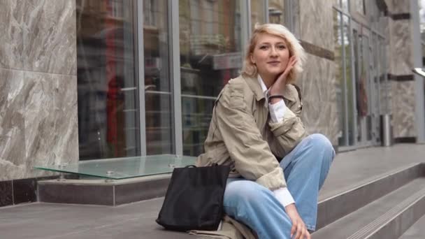 Молода блондинка в бежевому траншеї пальто, біла блузка і джинси сидять на сходах перед сучасною офісною будівлею — стокове відео
