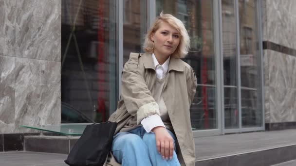 Ung blond kvinna i en beige trenchcoat, vit blus och jeans sitter på framtrapporna i en modern kontorsbyggnad och tittar på kameran — Stockvideo