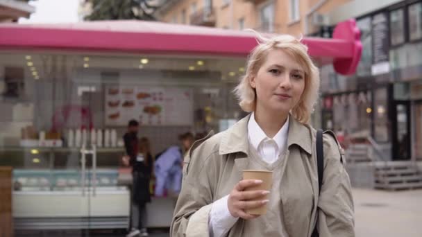 Joven mujer de negocios rubia en una gabardina beige y chaleco, blusa blanca se levanta con una taza de cartón desechable de café en el fondo de un café de la calle y mira a la cámara — Vídeos de Stock