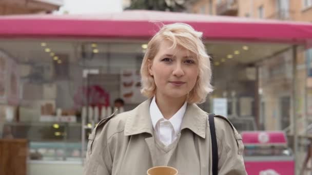 身穿米黄色风衣和背心的年轻金发女商人站在一家快餐店的后面，手里拿着一杯一次性纸杯咖啡，看着摄像机 — 图库视频影像