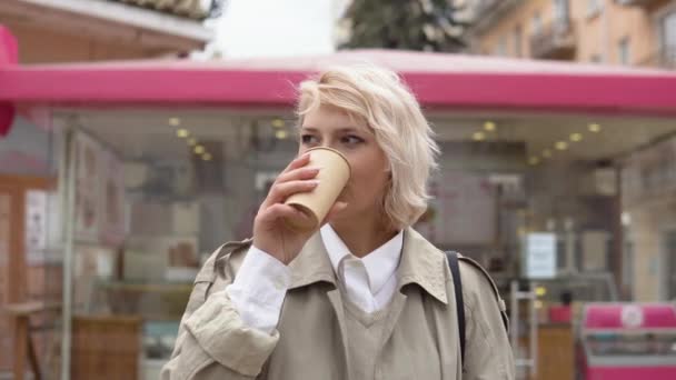 Mladé blondýny obchodní žena v béžové trenč kabát a vesta, bílá blůza pije kávu z kartonu jednorázové pohár na pozadí pouliční kavárny