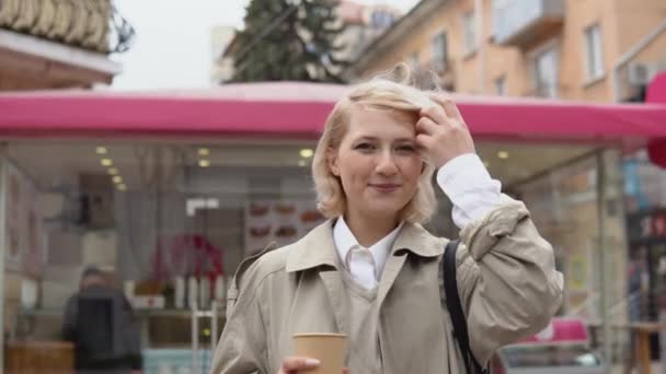 Bej renkli trençkotlu ve yelekli genç sarışın iş kadını, şehirde beyaz bir bluz kahve içer. — Stok video