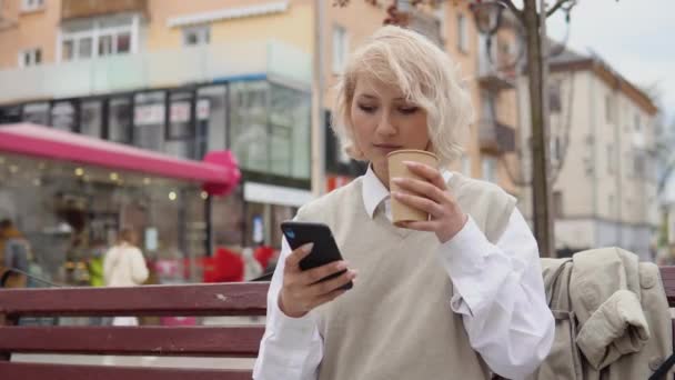 Junge blonde Geschäftsfrau in beiger Weste und weißer Bluse sitzt auf einer Bank inmitten der Stadt und arbeitet mit einem Imbisskaffee in der Hand in der Nähe des Straßencafés an einem Handy im Internet. — Stockvideo