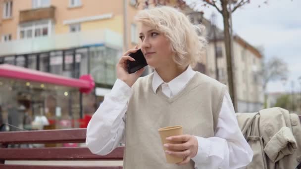 Młoda blondynka bizneswoman w beżowej kamizelce i białej bluzce siedzi na ławce w środku miasta i rozmawia przez telefon komórkowy online z kawą na wynos w ręku — Wideo stockowe